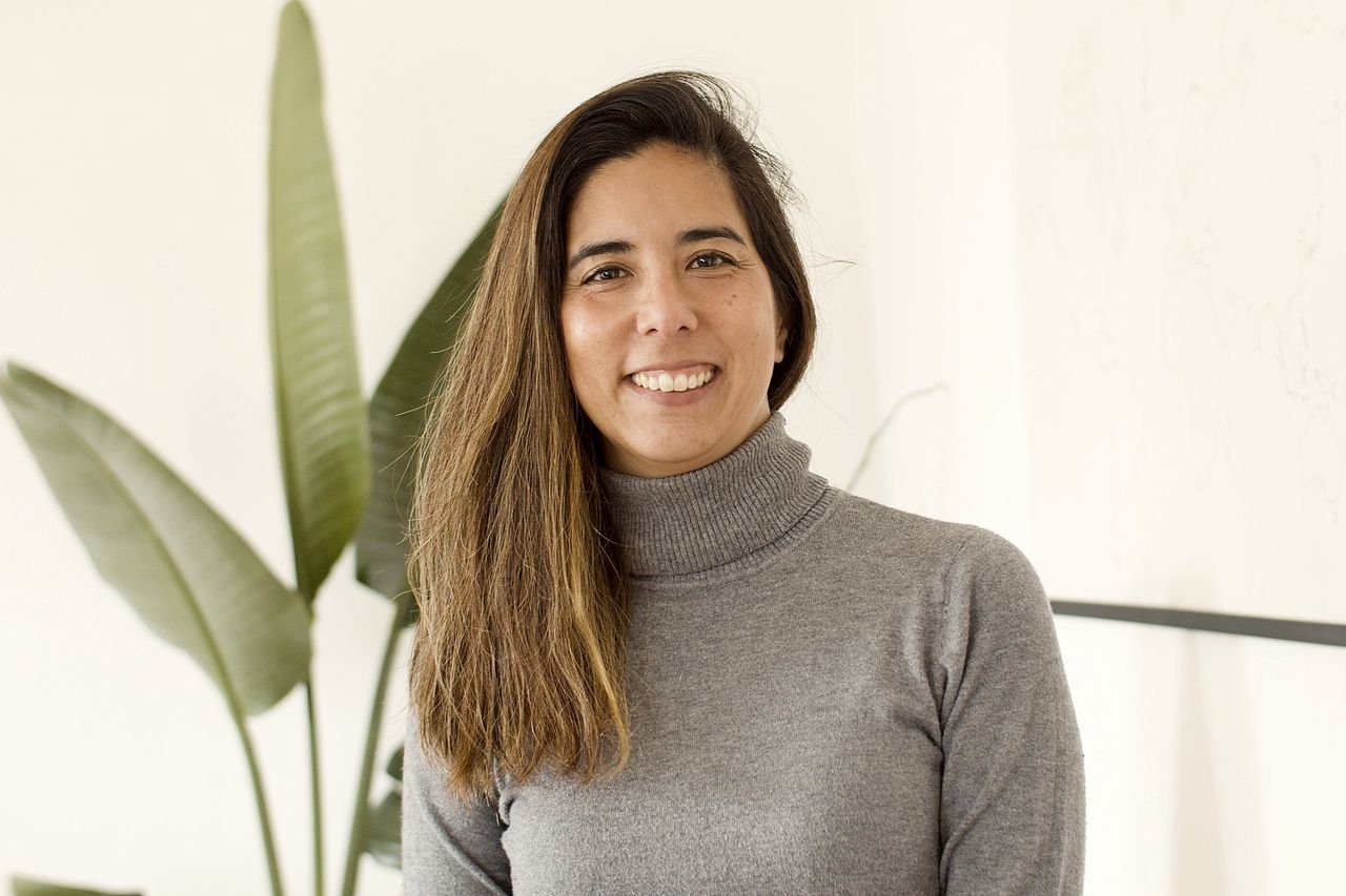 Portrait of a smiling woman in a gray turtleneck against a stylish indoor plant backdrop.